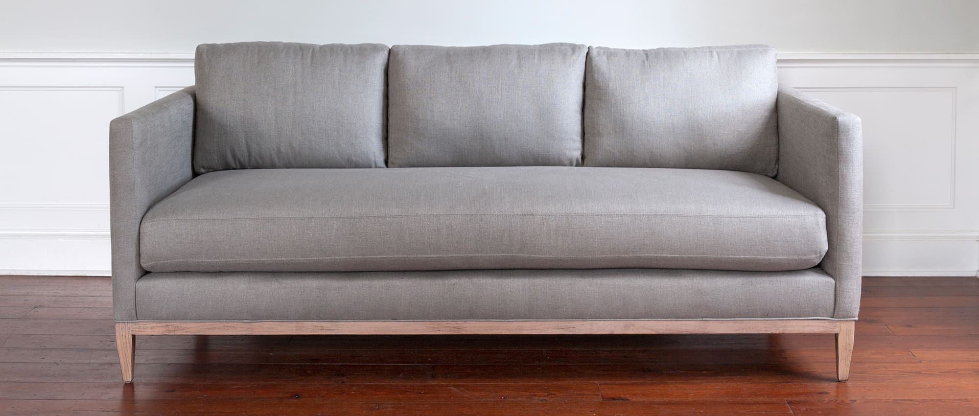 The Henry Sofa is pictured in medium grey, with an exposed wooden base, one long seat cushion, and three back cushions. Find it at GDC's Mount Pleasant furniture stores.